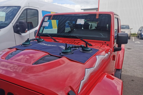 Remplacement de pare-brise en cours sur un 4x4 rouge chez Mondial Pare-Brise, avec outils de levage posés sur le capot pour une intervention sécurisée.