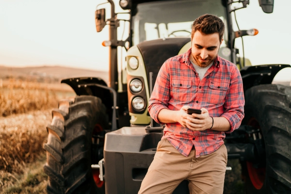 Jeune agriculteur appuyé contre son tracteur, consultant son smartphone en pleine nature, symbole d’une agriculture connectée et moderne.